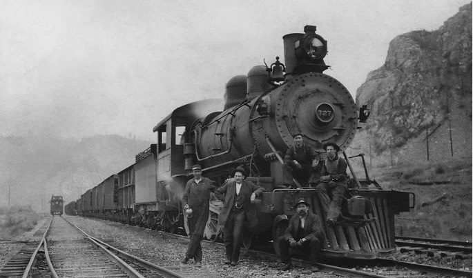 A black and white photo of men standing on the tracks next to an old train.